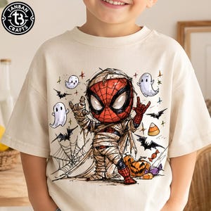 May include: A cream-colored short-sleeve t-shirt featuring a graphic illustration of a character dressed as a mummy with a red and black spider-web mask. The design includes small white ghosts, black bats, orange pumpkins, and candy corn.