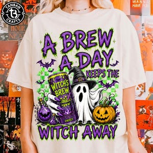 Puede incluir: Una camiseta color crema con un gráfico de un fantasma con sombrero de bruja, sosteniendo una lata etiquetada como Witch's Brew, junto a una calabaza y una poción. El texto dice A BREW A DAY KEEPS THE WITCH AWAY en letras moradas y verdes.