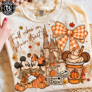 May include: A cream-colored t-shirt featuring a graphic print with the text "Fall vibes on Main street" and "autumn SEASON". The design includes a castle, Mickey and Minnie Mouse in scarves, a large orange gingham bow, and a pumpkin spice drink.
