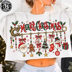 May include: A white long-sleeve cropped sweatshirt featuring a large graphic of a gold safety pin holding various Christmas ornaments including Santa Claus, a gingerbread man, a candy cane, a tree, a stocking, a snowman, and a gift box.