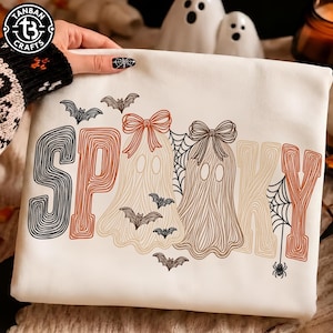 May include: A white sweatshirt featuring the word SPOOKY in a line-art style with orange, black, and tan colors. The design includes two ghosts wearing bows, several small flying bats, and spider webs with a hanging spider integrated into the letters.