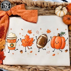 May include: A white sweatshirt featuring a colorful graphic of a smiling coffee cup, a maple leaf, a football, and a pumpkin with arms and legs. The design includes small orange leaves, acorns, and star accents. The logo TANBAN CRAFTS is in the corner.