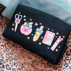 May include: A black t-shirt featuring a colourful graphic print of school supplies with faces. The design includes a pair of scissors, a pink apple, a yellow pencil, a pink notebook, and a ruler, all decorated with blue and pink bows and small hearts.