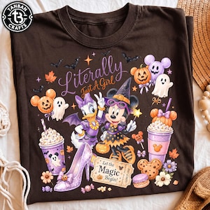 May include: A dark brown t-shirt featuring a Halloween-themed graphic with Daisy Duck and Minnie Mouse in costumes. The design includes pumpkins, ghosts, bats, a high-heeled shoe, a drink cup, a cookie, and text reading Literally Just A Girl and Let the Magic Begin!