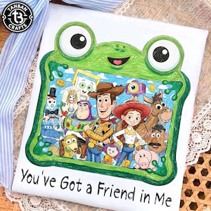 May include: A white t-shirt featuring a green frog-shaped frame illustration containing a group of cartoon characters including a cowboy, a cowgirl, a space ranger, and a horse. The text "You've Got a Friend in Me" is printed in black below the frame.