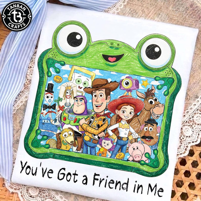 May include: A white t-shirt featuring a green frog-shaped frame illustration containing a group of cartoon characters including a cowboy, a cowgirl, a space ranger, and a horse. The text "You've Got a Friend in Me" is printed in black below the frame.