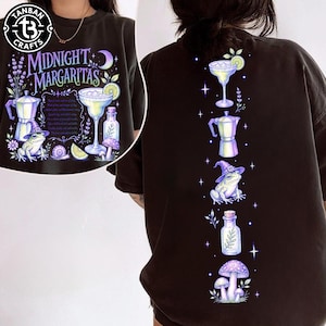 May include: A black t-shirt featuring a graphic print with the text Midnight Margaritas. The design includes a margarita glass, a moka pot, a frog wearing a witch hat, a glass bottle, a snail, mushrooms, and lavender in shades of purple and green.
