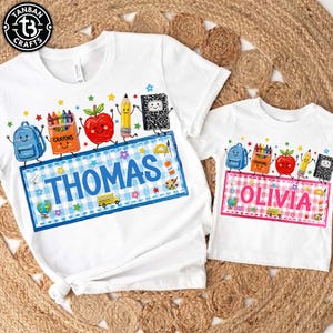 May include: Two white t-shirts featuring school-themed graphics including a backpack, crayons, apple, pencil, and notebook. The larger shirt displays the name THOMAS in blue, while the smaller shirt displays the name OLIVIA in pink.