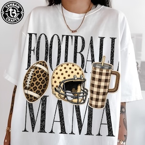 May include: A white t-shirt featuring the black text FOOTBALL MAMA. The design includes a leopard print football, a tan polka-dot football helmet, and a brown and tan buffalo plaid tumbler cup with a straw, all arranged across the center.