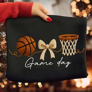 May include: Black decorative pillow featuring a basketball, a bow, and a basketball net design. The words "Game day" are written in white script. The basketball is brown and orange, and the net is beige.
