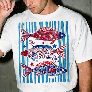 May include: White t-shirt featuring three fish designs in red, white, and blue, with star and stripe patterns. The fish are set against a backdrop of blue and gold vertical stripes, creating a patriotic theme.
