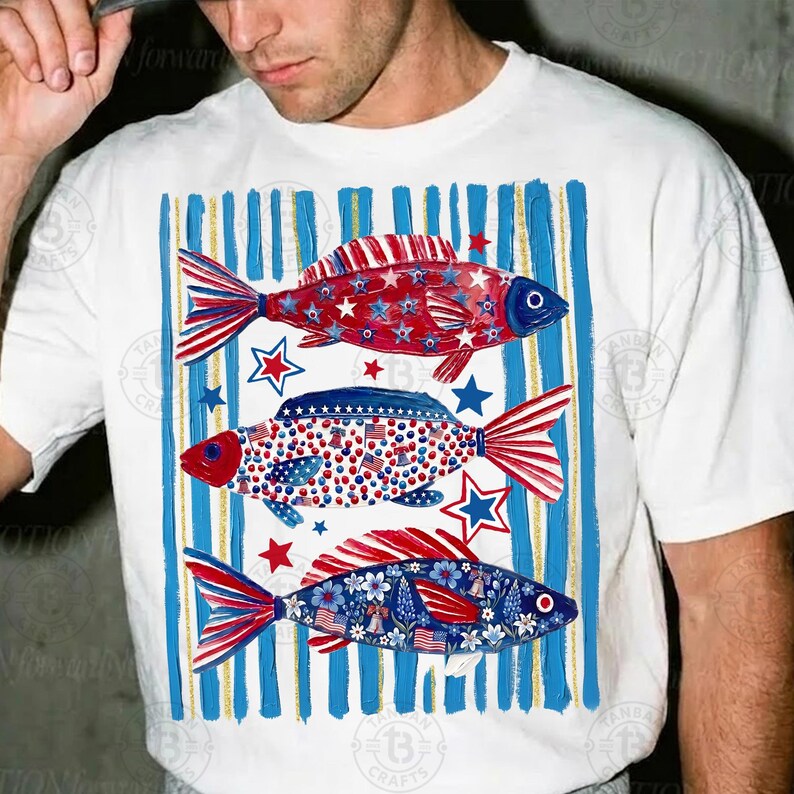 May include: White t-shirt featuring three fish designs in red, white, and blue, with star and stripe patterns. The fish are set against a backdrop of blue and gold vertical stripes, creating a patriotic theme.