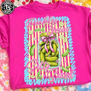 May include: A bright pink t-shirt featuring a jar of pickles with a pink bow, surrounded by pickle slices and the text "Pickles and Jesus Patty & Paved". The shirt has a round neck and short sleeves.