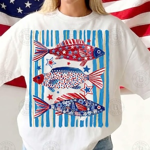 May include: White t-shirt featuring three fish designs in red, white, and blue. The fish are decorated with stars, stripes, and floral patterns, set against a background of blue and gold vertical stripes. The design evokes a patriotic theme.