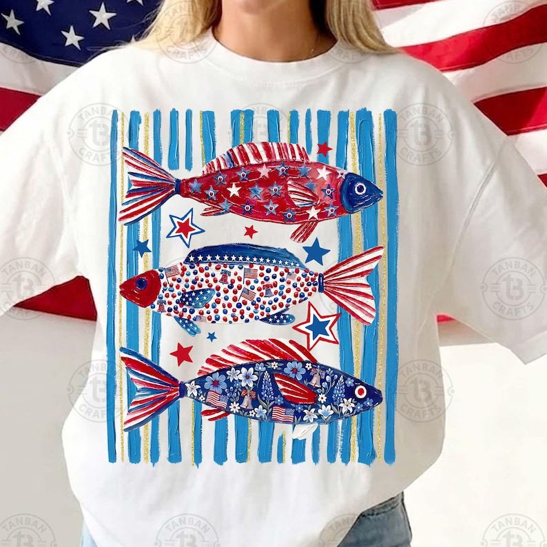 May include: White t-shirt featuring three fish designs in red, white, and blue. The fish are decorated with stars, stripes, and floral patterns, set against a background of blue and gold vertical stripes. The design evokes a patriotic theme.