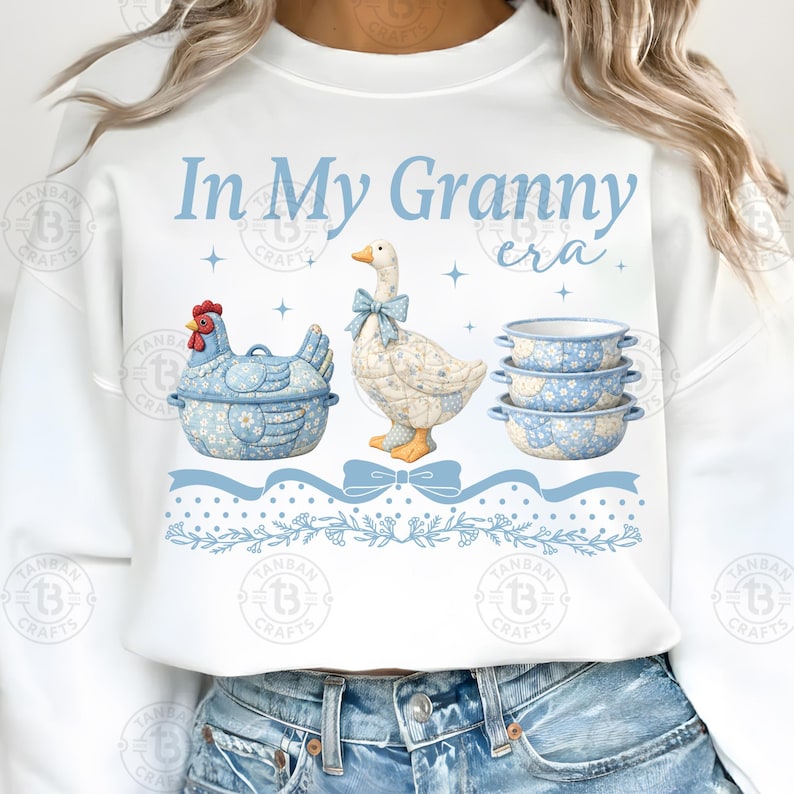 May include: White sweatshirt with a light blue graphic that reads "In My Granny era." The graphic features a blue chicken-shaped container, a goose with a blue bow, and three blue floral bowls. A blue ribbon and floral border complete the design.