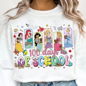 May include: White sweatshirt featuring a colorful graphic celebrating "100 days of school." The design includes illustrations of Disney princesses holding books, along with various school-themed elements like books, pencils, and a globe.