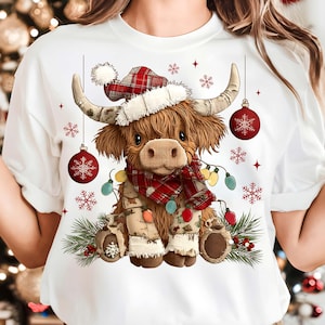 May include: White t-shirt featuring a Highland cow illustration. The cow wears a plaid hat, scarf, and festive lights, with hanging ornaments and snowflakes. The design has a Christmas theme.