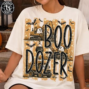 May include: A cream-colored t-shirt featuring a graphic print of construction vehicles, ghosts, bats, and pumpkins. The text "BOO DOZER" is printed in large black letters. The design includes orange and yellow accents with a polka dot pattern.