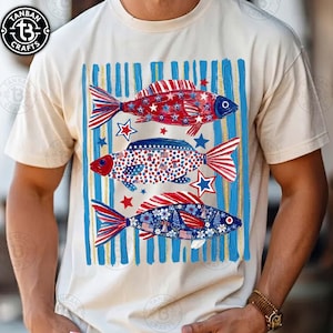 May include: Cream-colored t-shirt featuring three decorative fish in red, white, and blue. The fish are adorned with stars, stripes, and floral patterns, set against a backdrop of blue and gold vertical stripes. The shirt has a crew neck and short sleeves.