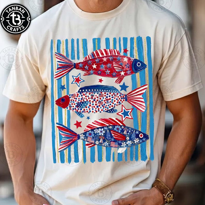May include: Cream-colored t-shirt featuring three decorative fish in red, white, and blue. The fish are adorned with stars, stripes, and floral patterns, set against a backdrop of blue and gold vertical stripes. The shirt has a crew neck and short sleeves.