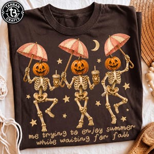 Pode incluir: Uma t-shirt castanho-escura com um gráfico de três esqueletos com cabeças de abóbora, cada um segurando um guarda-chuva às riscas e uma bebida. O design inclui estrelas amarelas, uma lua crescente e o texto: me trying to enjoy summer while waiting for fall.