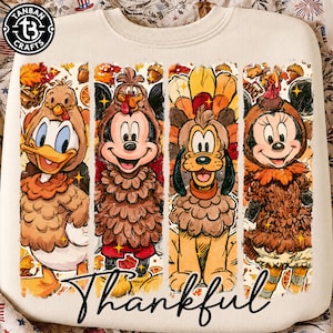 May include: A cream-colored crewneck sweatshirt featuring a graphic print of four cartoon characters dressed in turkey costumes. The design includes autumn leaves, acorns, and the word Thankful in black script. A Tanban Crafts logo is in the corner.