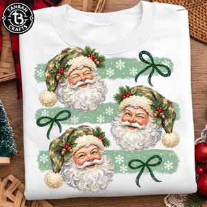 May include: A white sweatshirt featuring three illustrations of Santa Claus wearing camouflage-patterned hats adorned with holly berries and pine cones. The design includes light green horizontal brush strokes, white snowflakes, and dark green bows.