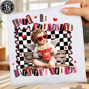 May include: White t-shirt with the text "LOVE IS OVERRATED" and "PASS THE SNACKS". The design features a cherub wearing heart-shaped sunglasses, holding a popcorn bucket, and sitting on a bench with a black and white checkered background.