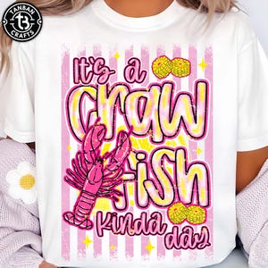 May include: White t-shirt featuring a pink and yellow graphic. The design reads "It's a Crawfish Kinda Day" with a pink crawfish illustration. The graphic includes yellow stars and flower accents, set against pink and white vertical stripes.