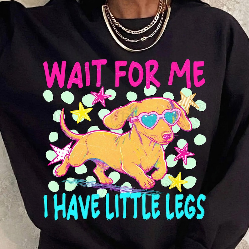 May include: Black sweatshirt with a colorful graphic of a dachshund wearing heart-shaped sunglasses. The text reads "WAIT FOR ME I HAVE LITTLE LEGS" in pink and blue. The design includes stars and polka dots.