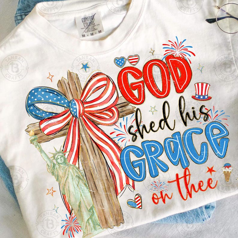 May include: Cream-colored t-shirt with a patriotic design. Features a wooden cross adorned with a red, white, and blue bow, the Statue of Liberty, and the text "GOD shed his GRACE on thee."