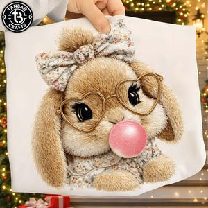 Op de afbeelding: Een wit kussen met een gedetailleerde illustratie van een bruin konijn met hartvormige bril en een roze kauwgomballenbel. Het konijn draagt een bloemenstrik en een trui. Het kussen wordt door een hand vastgehouden.