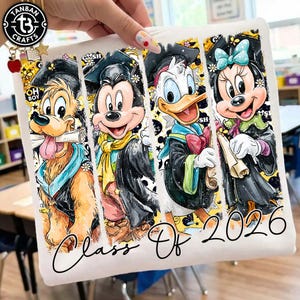 May include: White square fabric featuring four cartoon characters: Goofy, Mickey Mouse, Donald Duck, and Minnie Mouse, all in graduation attire. The words "Class Of 2026" are written in cursive below the characters. The fabric is held by a hand.