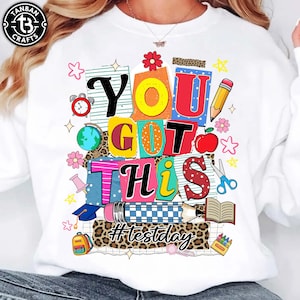 May include: White sweatshirt with a colorful graphic that says "YOU GOT THIS" in various fonts and colors. The design includes school-themed elements like a pencil, apple, and scissors. The hashtag "#testday" is also included.
