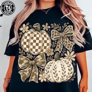 May include: A black short-sleeve t-shirt featuring a graphic print of two pumpkins, one with a brown and white checkered pattern and one with a white leaf design, accented by a leopard print bow, a camouflage bow, and small flowers.