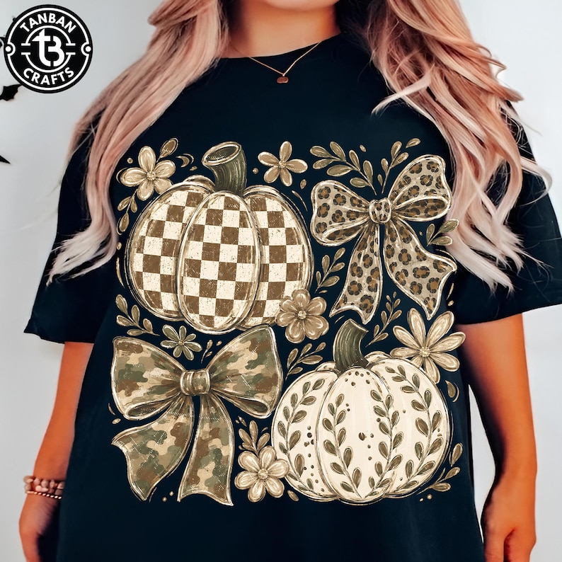 May include: A black short-sleeve t-shirt featuring a graphic print of two pumpkins, one with a brown and white checkered pattern and one with a white leaf design, accented by a leopard print bow, a camouflage bow, and small flowers.