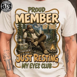 Pode incluir: Camiseta bege com um gráfico de Bigfoot relaxando em uma cena de floresta, segurando uma caneca. O texto diz "PROUD MEMBER OF THE JUST RESTING MY EYES CLUB." O design inclui uma fogueira, montanhas e o texto "A DAD LIKE ME IS HARD TO FIND."