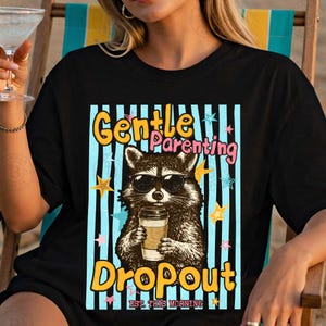 May include: Black t-shirt featuring a raccoon wearing sunglasses and holding a coffee cup. The shirt has the words "Gentle Parenting Dropout" in yellow and pink, with a blue striped background and star accents.