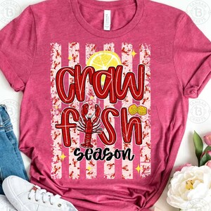 May include: A red t-shirt with the words "Crawfish Season" in a bold, stylized font. The design includes a crawfish illustration, a lemon slice, and peanuts. The background features red and white stripes with crawfish and star accents.