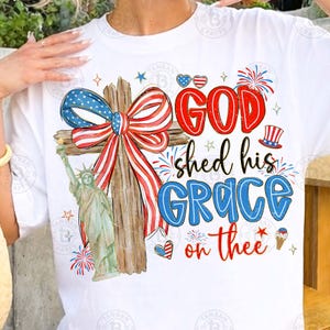 May include: White t-shirt featuring a patriotic design with a wooden cross adorned with a red, white, and blue bow. The shirt includes the text "GOD shed his GRACE on thee" with illustrations of fireworks, hearts, and the Statue of Liberty.