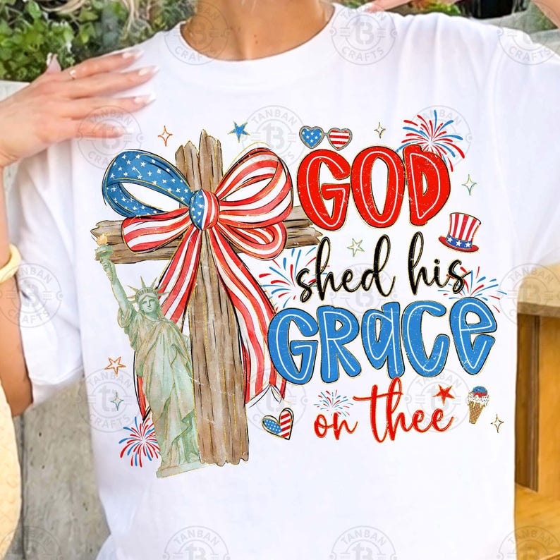 May include: White t-shirt featuring a patriotic design with a wooden cross adorned with a red, white, and blue bow. The shirt includes the text "GOD shed his GRACE on thee" with illustrations of fireworks, hearts, and the Statue of Liberty.