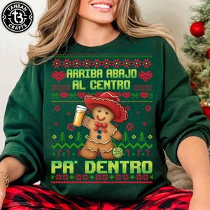 May include: Green sweatshirt with a gingerbread man wearing a red hat and holding a beer. The sweatshirt has the text "ARRIBA ABAJO AL CENTRO PA' DENTRO" in white and green. The sweatshirt has a festive, holiday theme.