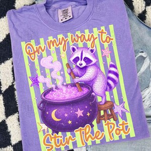 May include: A light purple t-shirt featuring a graphic of a raccoon stirring a purple cauldron with a wooden spoon. The design includes light green vertical stripes, yellow stars, a crescent moon, and the text On my way to Stir The Pot.