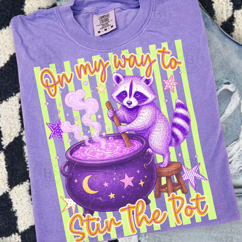 May include: A light purple t-shirt featuring a graphic of a raccoon stirring a purple cauldron with a wooden spoon. The design includes light green vertical stripes, yellow stars, a crescent moon, and the text On my way to Stir The Pot.