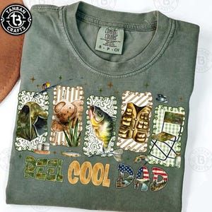 Op de afbeelding: Olijfgroen T-shirt met een visserij-thema grafisch ontwerp. Het ontwerp bevat illustraties van visgerei, een vis en de tekst "Reel Cool Dad" in verschillende lettertypen en kleuren. Het shirt heeft een ronde hals en korte mouwen.