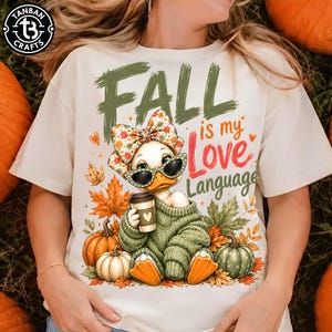 May include: A cream-colored t-shirt featuring a graphic of a duck wearing a floral bow and sunglasses, holding a coffee cup while wearing a green sweater. The text reads "FALL is my Love Language" surrounded by orange and green autumn leaves and pumpkins.