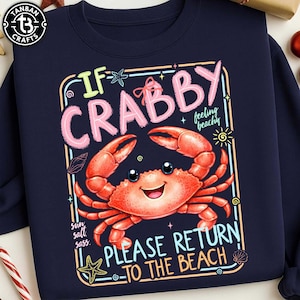 Pode incluir: Camisola azul marinho com um desenho de caranguejo de desenho animado. O desenho inclui o texto "If Crabby, Please Return to the Beach" em uma fonte divertida, juntamente com um caranguejo sorridente, estrelas do mar e conchas.