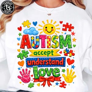 以下が含まれることがあります： カラフルなグラフィックデザインの白いスウェットシャツ。デザインには、赤、青、緑色の「自閉症」という言葉、パズルのピース、ハート、星、そして「受け入れる、理解する、愛する」という言葉が含まれています。