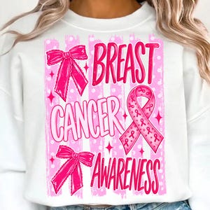 May include: A white crewneck sweatshirt featuring a graphic print with pink vertical stripes and polka dots. The design includes the text "BREAST CANCER AWARENESS" in pink, accompanied by two pink bows and a pink awareness ribbon with small ribbon icons.